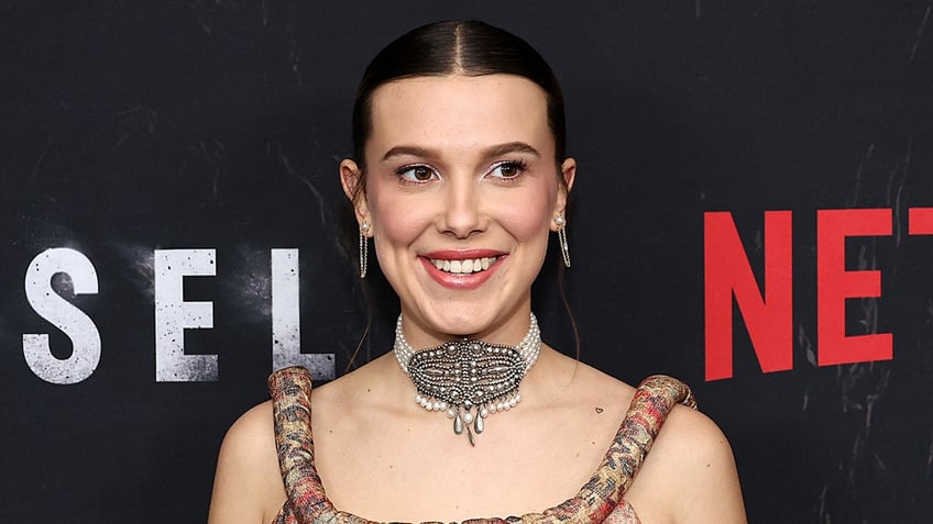 Millie Bobby Brown smiles and looks to her side as she stands on the carpet in a patterned tank top and choker