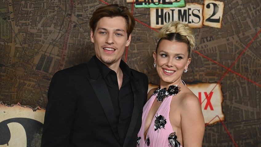 Jake Bon Jovi in a black suit and shirt smiles on the carpet with Millie Bobby Brown in a pink gown with dark flowers