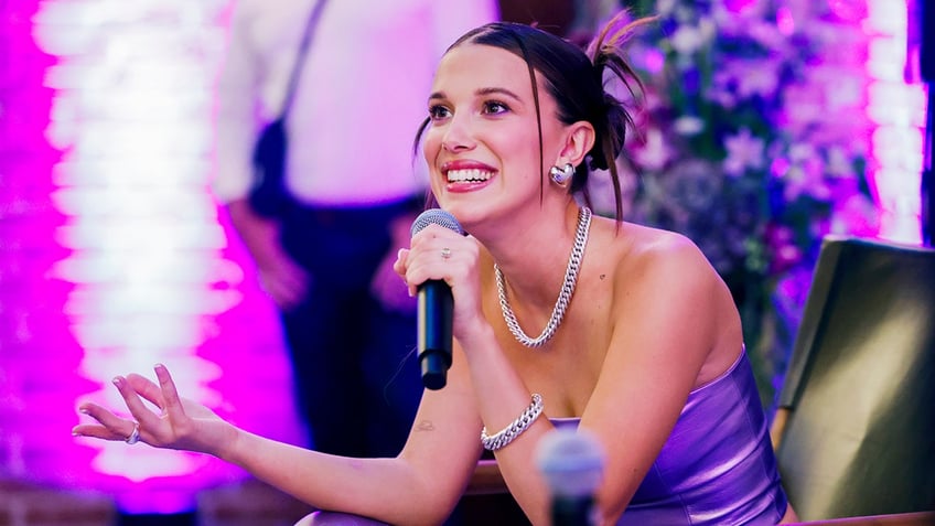 Millie Bobby Brown smiles on stage in a purple tube top as she holds the microphone to her mouth