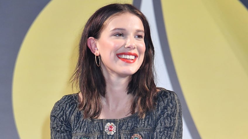 Millie Bobby Brown in a black jacket with colorful flower smiles on stage and looks to the side