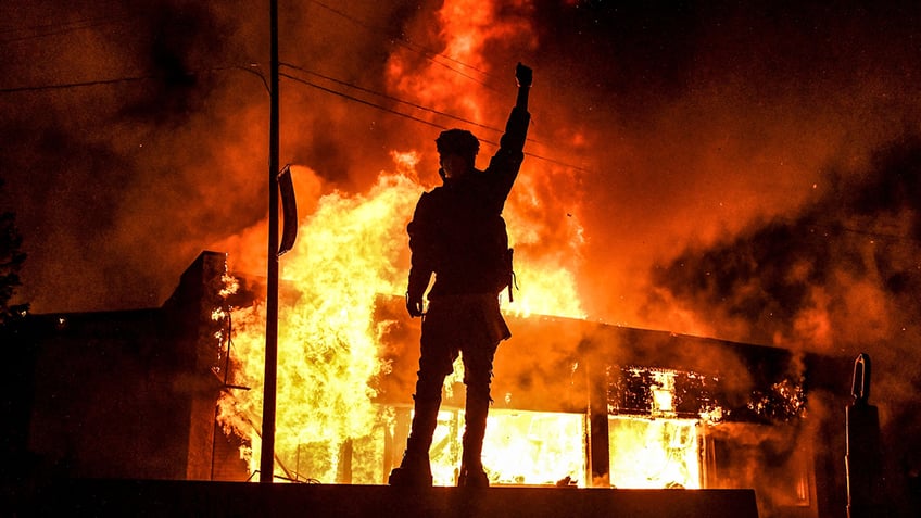 building on fire in background as defiant man raises arm in foreground