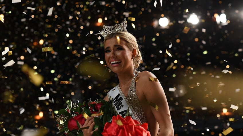 Abbie Stockward looks stunned holding a bouquet of red roses and wearing a crown as gold confetti falls down on her.