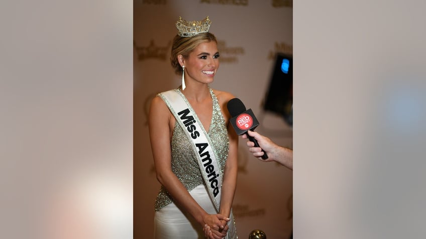 Abbie Stockard wearing a silver sparkling gown with her crown and a sash being interviewed.