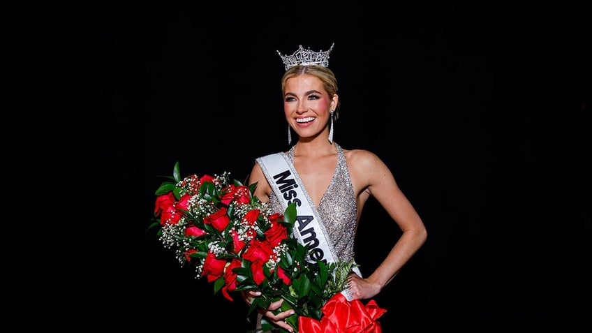 Miss America smiling and posing in a silver sparkling gown and holding a bouquet of red roses wearing a crown.