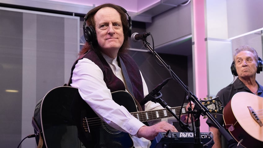 Tommy James sitting in a studio with headphone on and holding a guitar