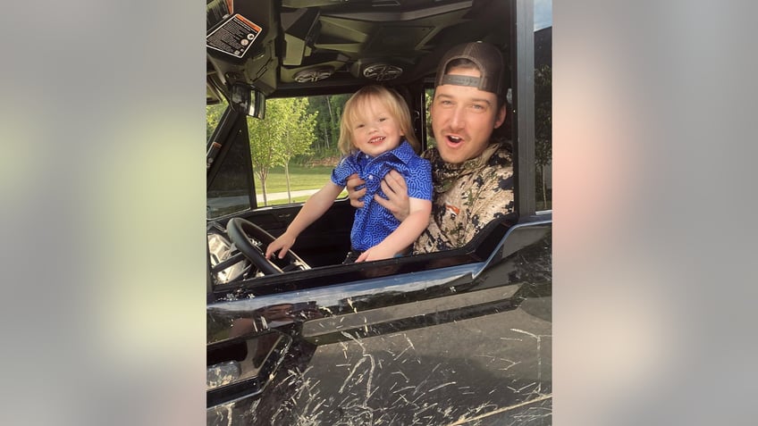 Morgan Wallen in a camo shirt and backwards hat holds his son in a blue shirt in his car