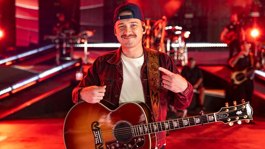 Morgan Wallen in a cream shirt and red jean jacket points to himself on stage at the BBMAs