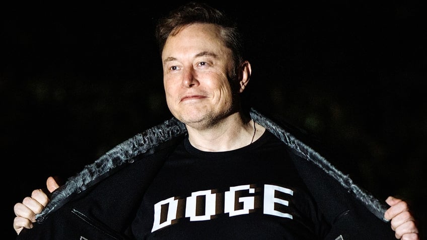 Elon Musk smiles while displaying his "DOGE" shirt