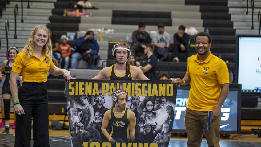 Siena Palmisciano stands with a poster celebrating her 100 wins wrestling. Her coaches Rebecca Mathewson and David Cureton stand with her.