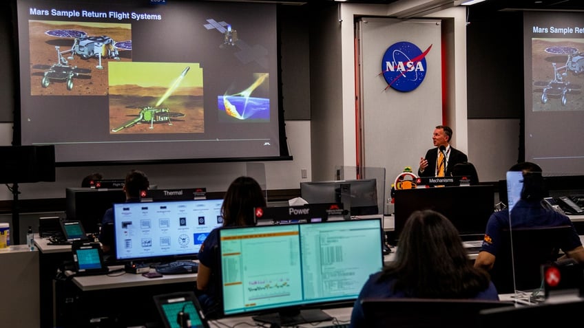 Bobby Braun gives a presentation on Mars sample return flight systems in Mars 2020 Perseverance Rover Mission Operations area of the Jet Propulsion Laboratory, filled with desks and computers.