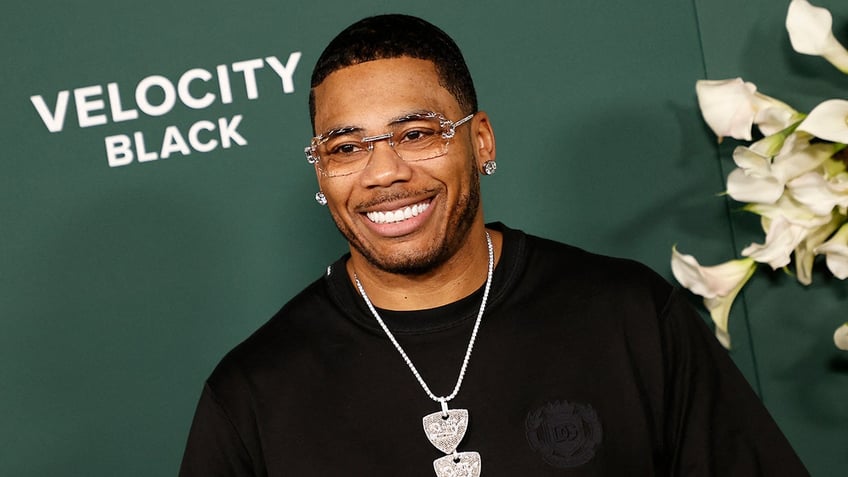 Nelly smiles in a black t-shirt with massive diamond earrings and a silver chain on the carpet, wearing clear eyeglasses