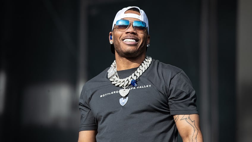 Nelly looks up on stage wearing a black t-shirt, a huge silver chain, backwards hat and blue-tinted sunglasses