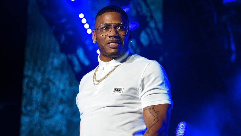 Nelly in a white polo shirt and chain necklace with transparent eyeglasses looks to his left on stage