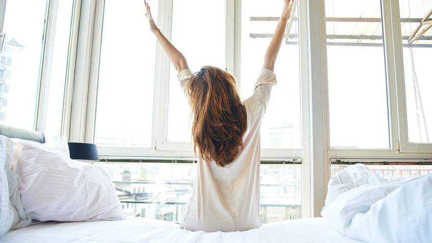 Young woman doing morning stretching in bed, arms raised, rear view