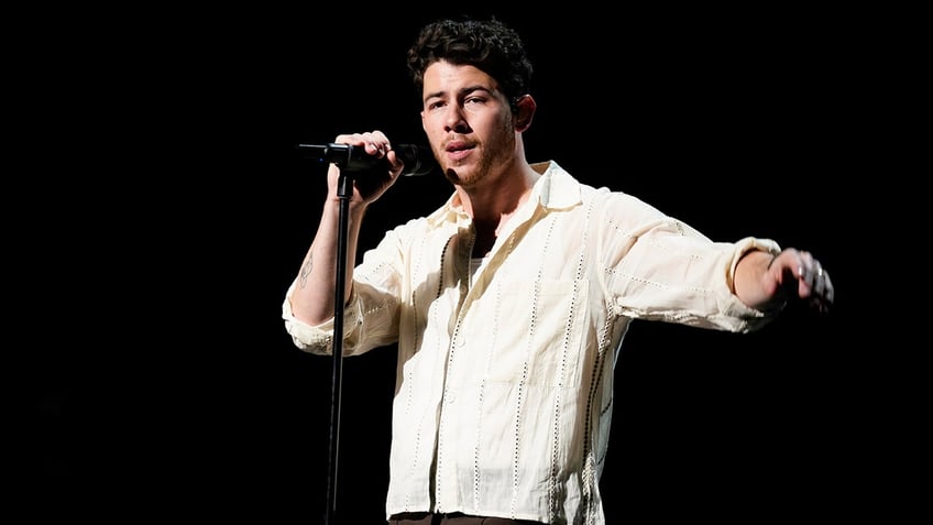 Nick Jonas outstretches his arm on stage holding the microphone with his other hand in a light yellow shirt