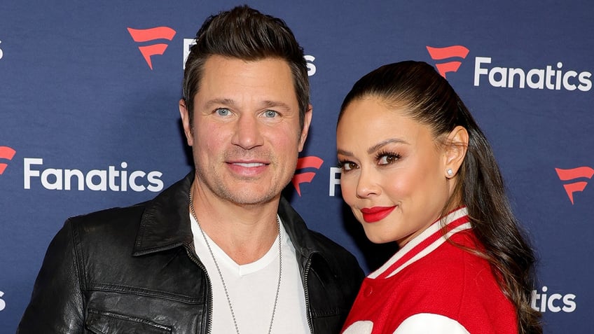 Nick Lachey in a leather jacket smiles on the carpet with wife Vanessa Lachey in a red letterman jacket