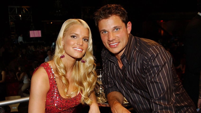 Jessica Simpson smiles wearing a sparkly red dress next to Nick Lachey in a silky black shirt with thin stripes