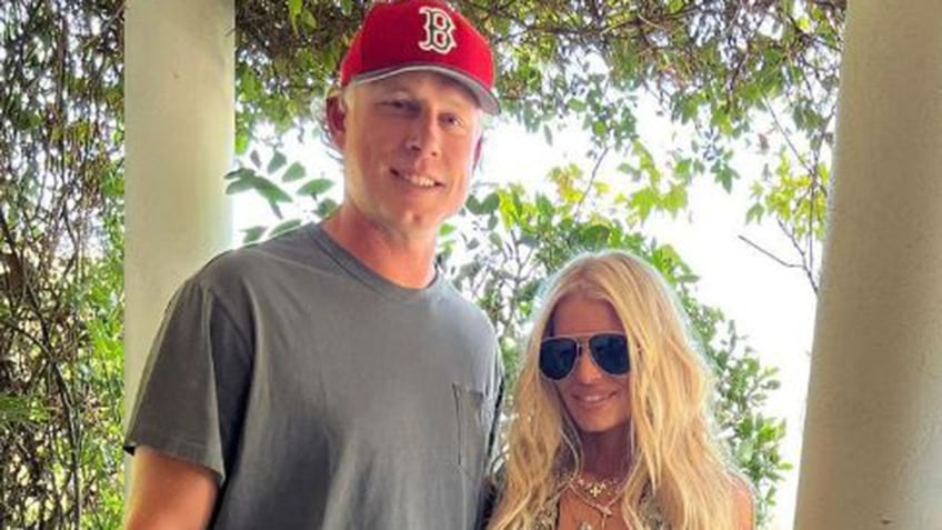 Eric Johnson in a grey shit and red Red Sox hat smiles next to wife Jessica Simpson