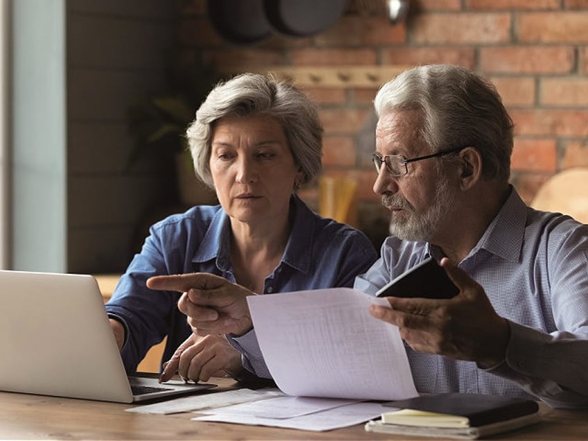 Focused senior husband and wife sit at table at home look at laptop screen pay bills taxes