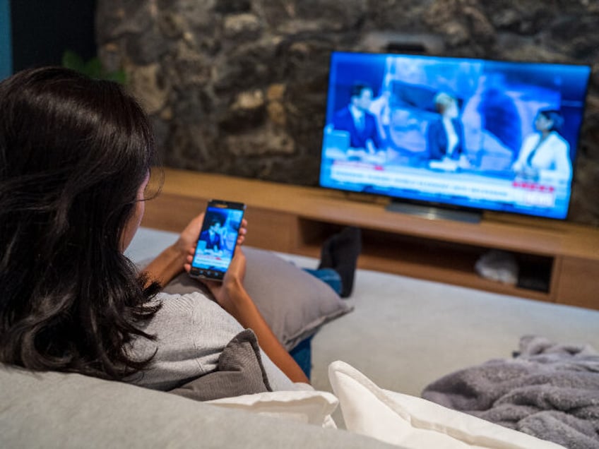 Rear view of young woman watching news on television and smart phone while sitting on sofa