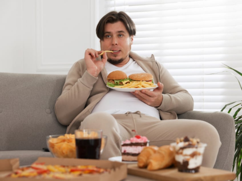 Overweight man with plate of burgers and French fries on sofa at home (Stock photo via Getty)