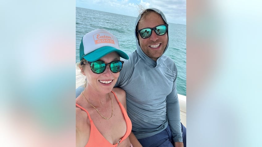India Oxenberg smiling in an organge bikini next to her husband in a light blue hoodie