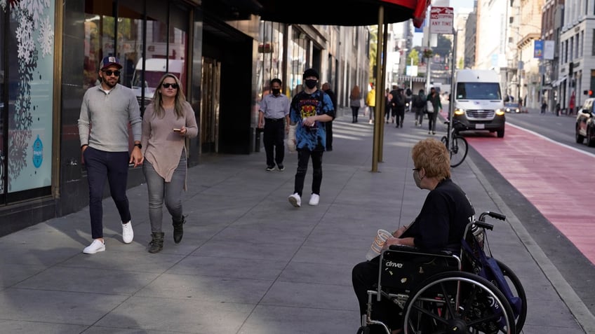 People pass by a woman in a wheelchair panhandling near Union Square in San Francisco, Thursday, Dec. 2, 2021. In San Francisco, homeless tents, open drug use, home break-ins and dirty streets have proliferated during the pandemic. (AP Photo/Eric Risberg)