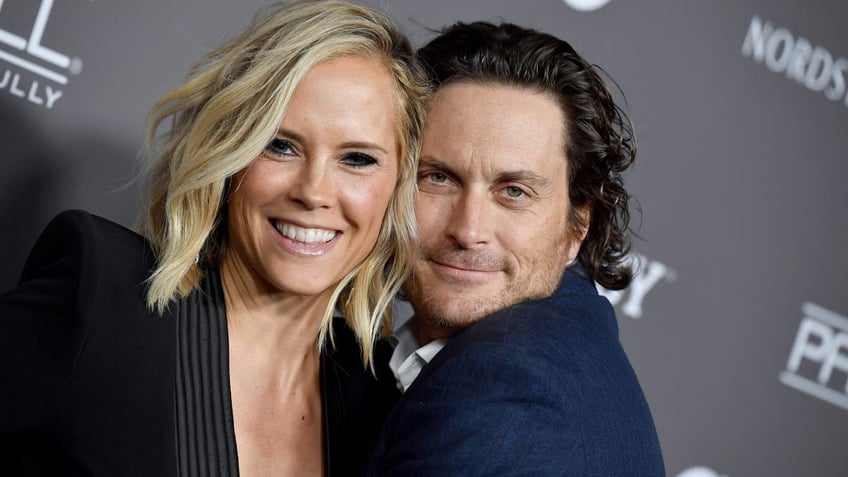 Oliver Hudson and Erin Bartlett hug and smile on carpet. 