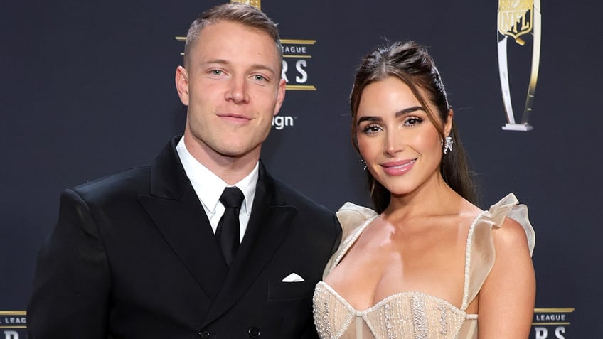 Christian McCaffrey in a black suit and tie soft smiles on the carpet with Olivai Culpo in a nude dress