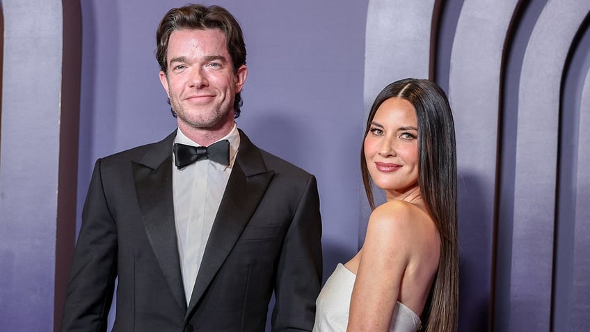 John Mulaney soft smiles at the Governors Awards in a classic tuxedo along with Olivia Munn in a white strapless dress slightly looking over her shoulder