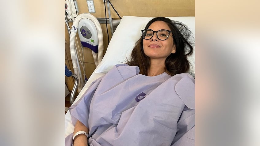 Olivia Munn soft smiles as she lays back in a hospital bed wearing a purple gown with black framed eye glasses