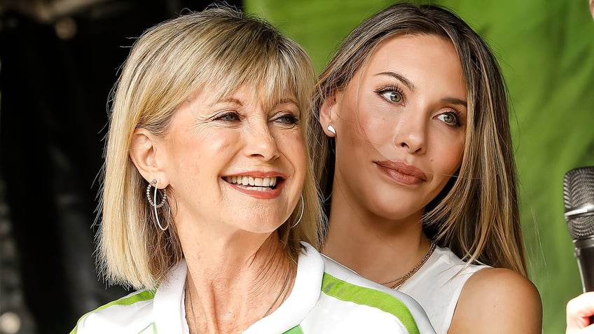 Olivia Newton-John smiles in a white top with green trim and silver hoops eras with her daughter Chloe hugging her from behind