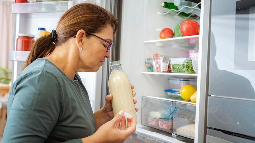 Disgusted woman smelling expired milk by the fridge.