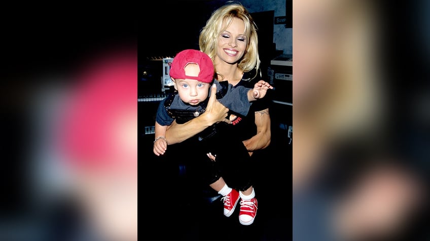 Pamela Anderson smiles and looks down at her son Brandon wearing red shoes and a red hat