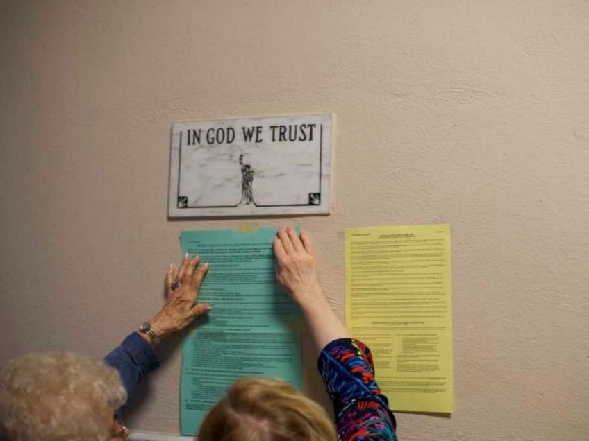 Election officials affix voting notices to the wall, below a plaque that states 'IN GOD WE