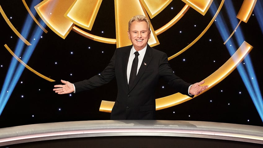 Pat Sajak in a black suit extends his arms on either side of him on set