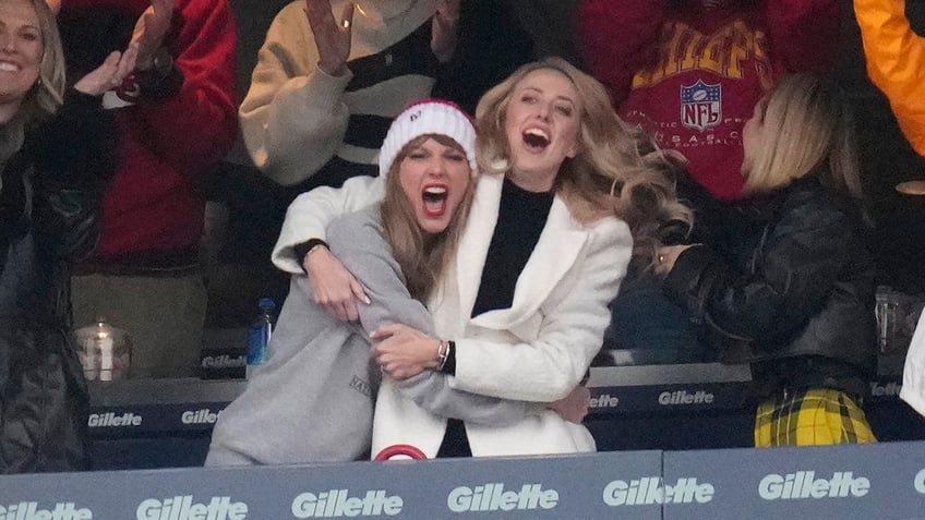 Taylor Swift in a grey sweatshirt excitedly hugs Brittany Mahomes in a white jacket