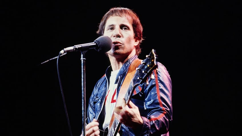 Singer Paul Simon wears a blue jacket and holds a guitar on stage.