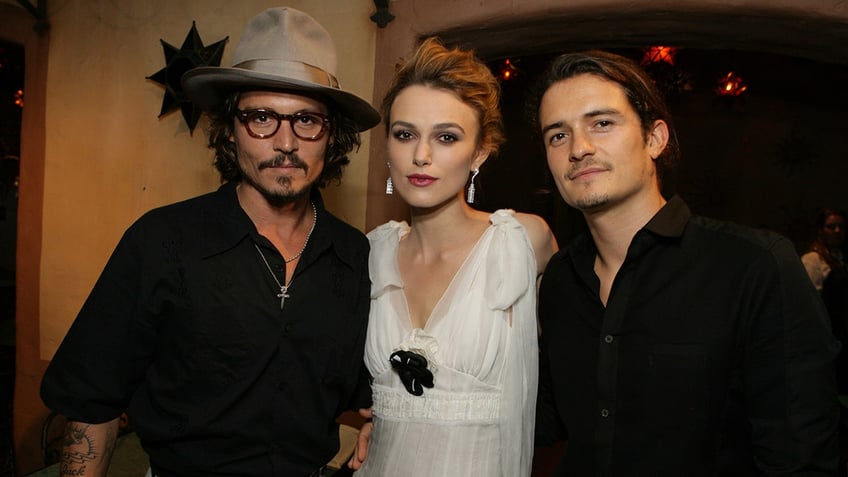 Johnny Depp in a black shirt and brown hat poses for a photo with Keira Knightley in a white dress and Orlando Bloom in a black shirt