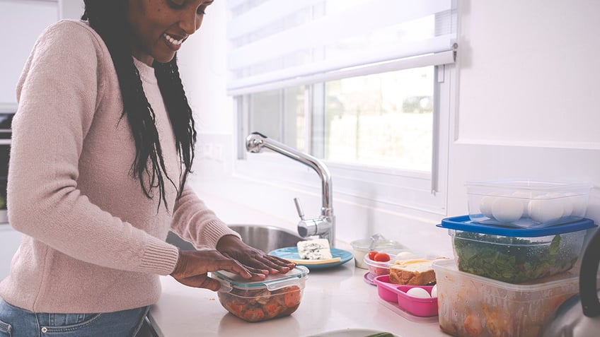 A young woman packing leftovers into a plastic container, cooking in a kitchen.