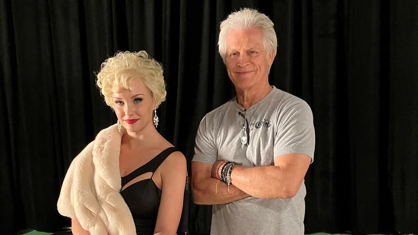 An actress posing as Stella Stevens sitting and wearing a black dress with a fur coat next to Andrew Stevens with his arms crossed wearing a grey shirt