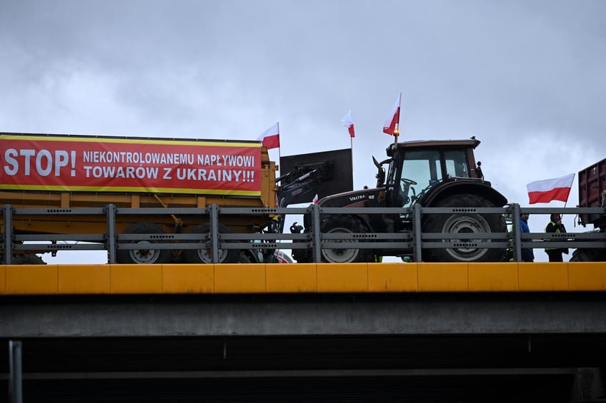 A banner reading 'Stop to uncontrolled supply of goods from Ukraine' is seen on a tractor trailer as a man carrying a Polish flag passes by while Polish farmers block the highway linking Warsaw and Lublin outside the town of Ryki, Lublin region, during a protest of farmers across the country againts EU climate measures on February 20, 2024. Farmers in Poland resumed a blockade of around 100 roads to the Ukrainian border in the morning of February 20 to protest against "uncontrolled" imports and demand a change to EU agricultural policy. For weeks Polish farmers have been blocking Ukrainian goods lorries from entering their country in anger at what they say is unfair competition from cheaper imports from Poland's war-torn neighbour and ally. (Photo by Sergei GAPON / AFP) (Photo by SERGEI GAPON/AFP via Getty Images)