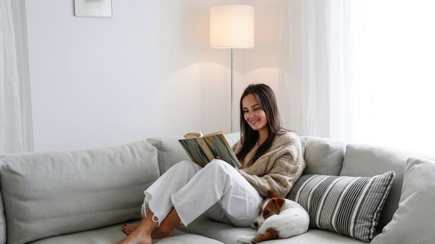 A woman reading a book on her couch at home with a dog by her side