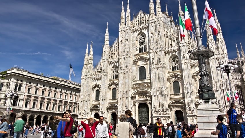 Milan Cathedral facade with flags on blue sky. The facade of the cathedral with many people walking. Flags waving on the blue sky.