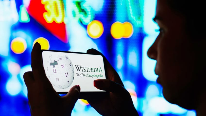 In this photo illustration, a silhouetted woman holds a smartphone with the Wikipedia logo displayed on the screen.