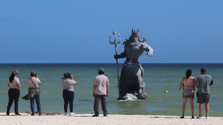 A stone statue of the Greek god Poseidon rises out of the ocean in Mexico as tourists snap photos.