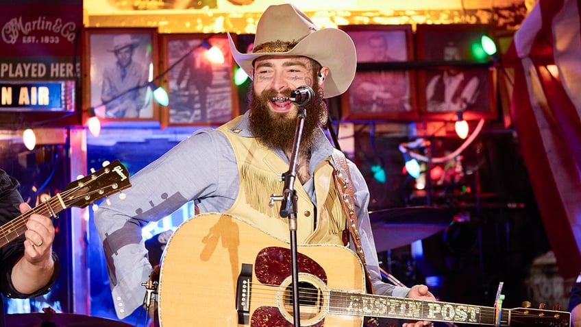 Post Malone in a yellow vest and blue shirt smiles while on stage in a tan cowboy hat