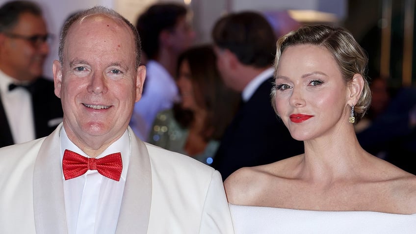 Prince Albert wearing a white suit with a red bow tie standing next to Princess Charlene wearing an off the shoulder white dress and red lipstick.