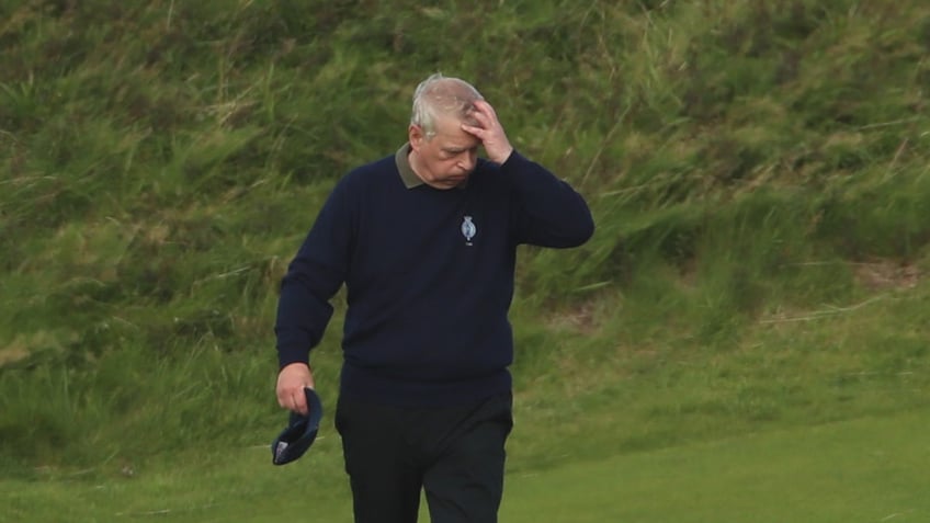 Prince Andrew looking stressed wearing a navy sweater and matching pants on a golf course.