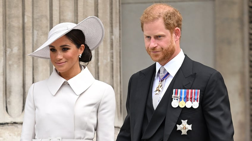 Meghan Markle wearing a white coatdress and a matching hat walking alongside Prince Harry in a suit with medals.